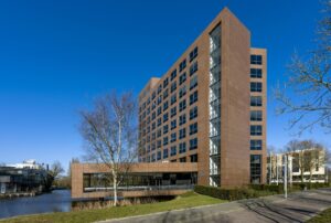 Modern kantoorgebouw aan de Prof. W.H. Keesomlaan met roodbruine gevel, grote ramen en een waterpartij op de voorgrond.